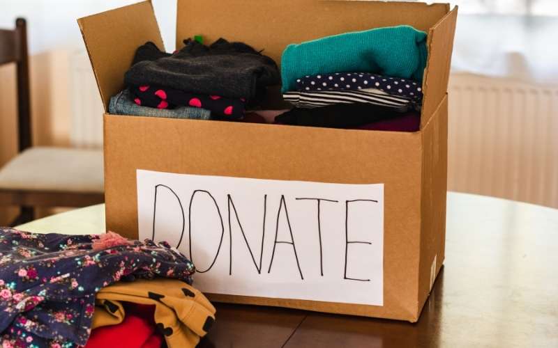 brown moving box with white piece of paper stuck on the front and the word "Donate" written on it, clothes piled up next to the box.