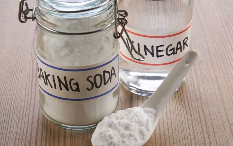 Image of a bottle of white vinegar, a jar of baking soda and a spoon with white powder on a brown table