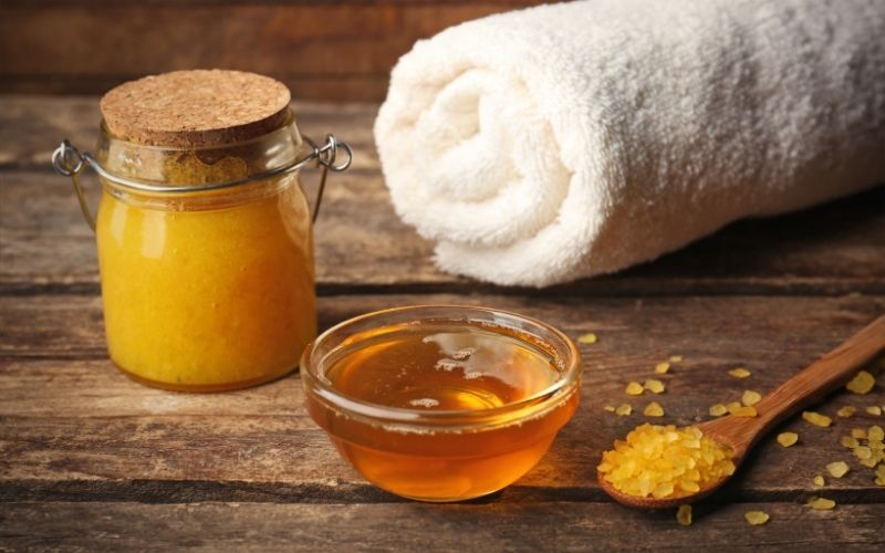 Photo of a natural scrub in a small jar, a bowl of honey a spoon of salt and a roll of white towel