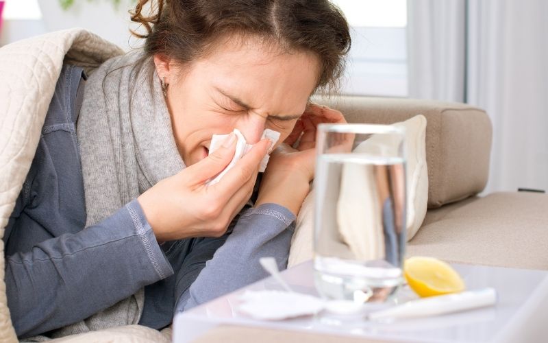 Phot of a woman holding her nose while in the bed with a glass of water near her.