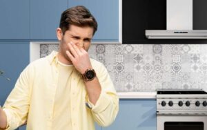 Photo of a man inside a house kitchen covering half of his face as if he smells something bad.