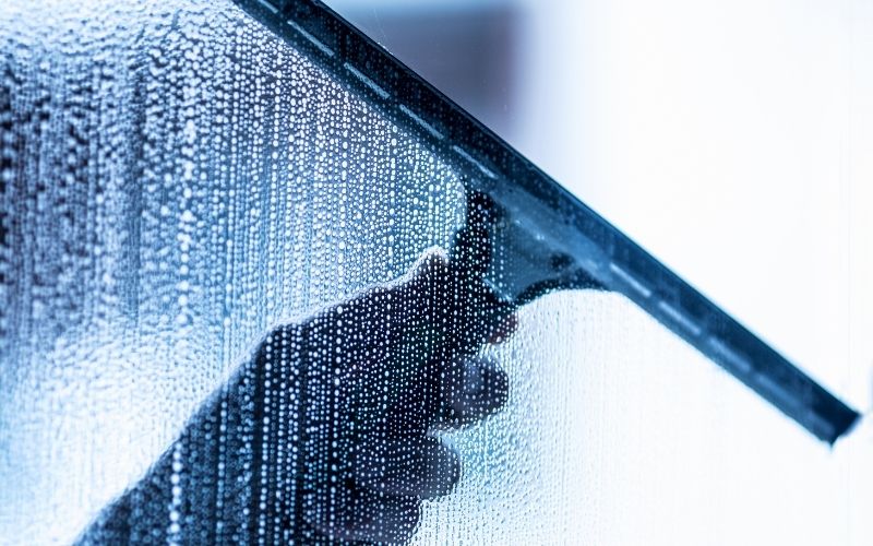 A hand, holding a squeegee while cleaning a shower glass