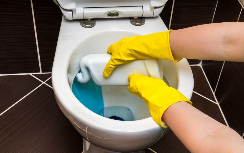 A pair of hands with yellow gloves holding a toilet cleaner squirting blue liquid into the bowl for cleaning